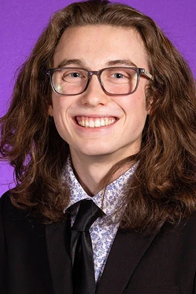 Zasha Griffin smiles in front of a purple painting. He has long, curly, brown hair and wears glasses, a bright purple floral shirt, a black tie and a black suit jacket.